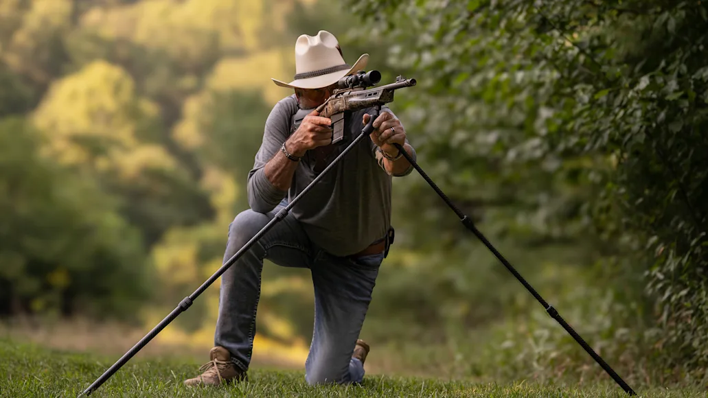 A shooter fires a rifle from the kneeling position, using shooting sticks for support.