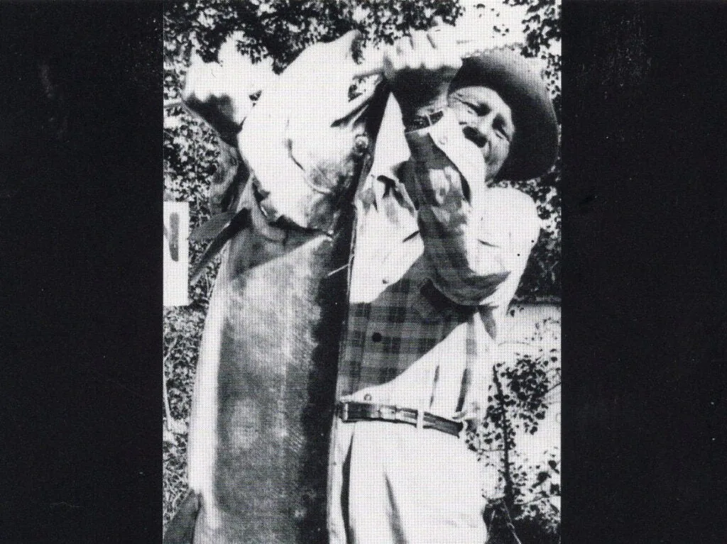 A black and white photo of a man holding up a large muskie fish.