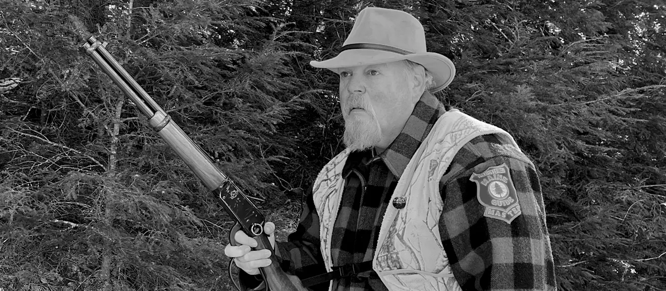 A hunter wearing blaze or and check jacket walks through the woods carrying a lever-action rifle. 