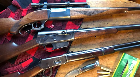 Three classic deer rifles sitting on a table with a wool jacket, knife, and ammo. 