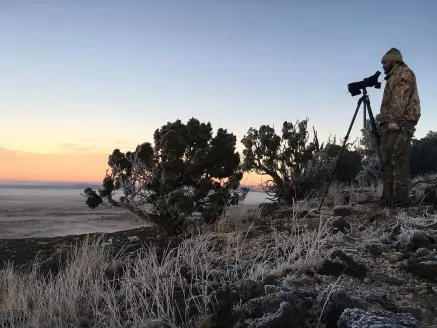 A hunter in camo scouts a western countryside with a spotting scope.