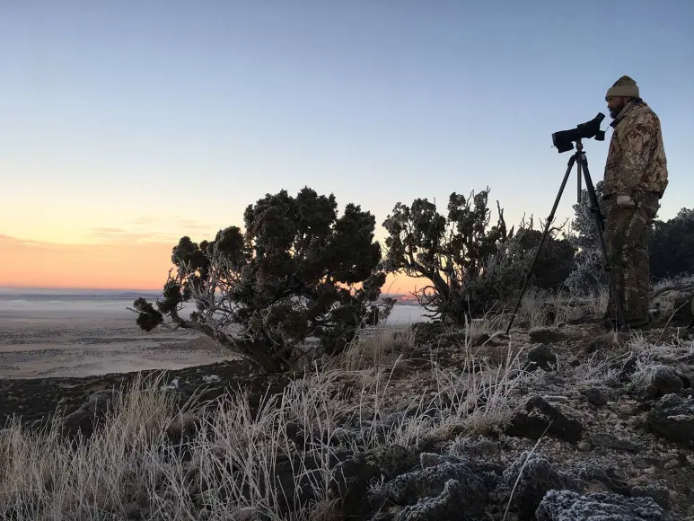 A hunter in camo scouts a western countryside with a spotting scope.