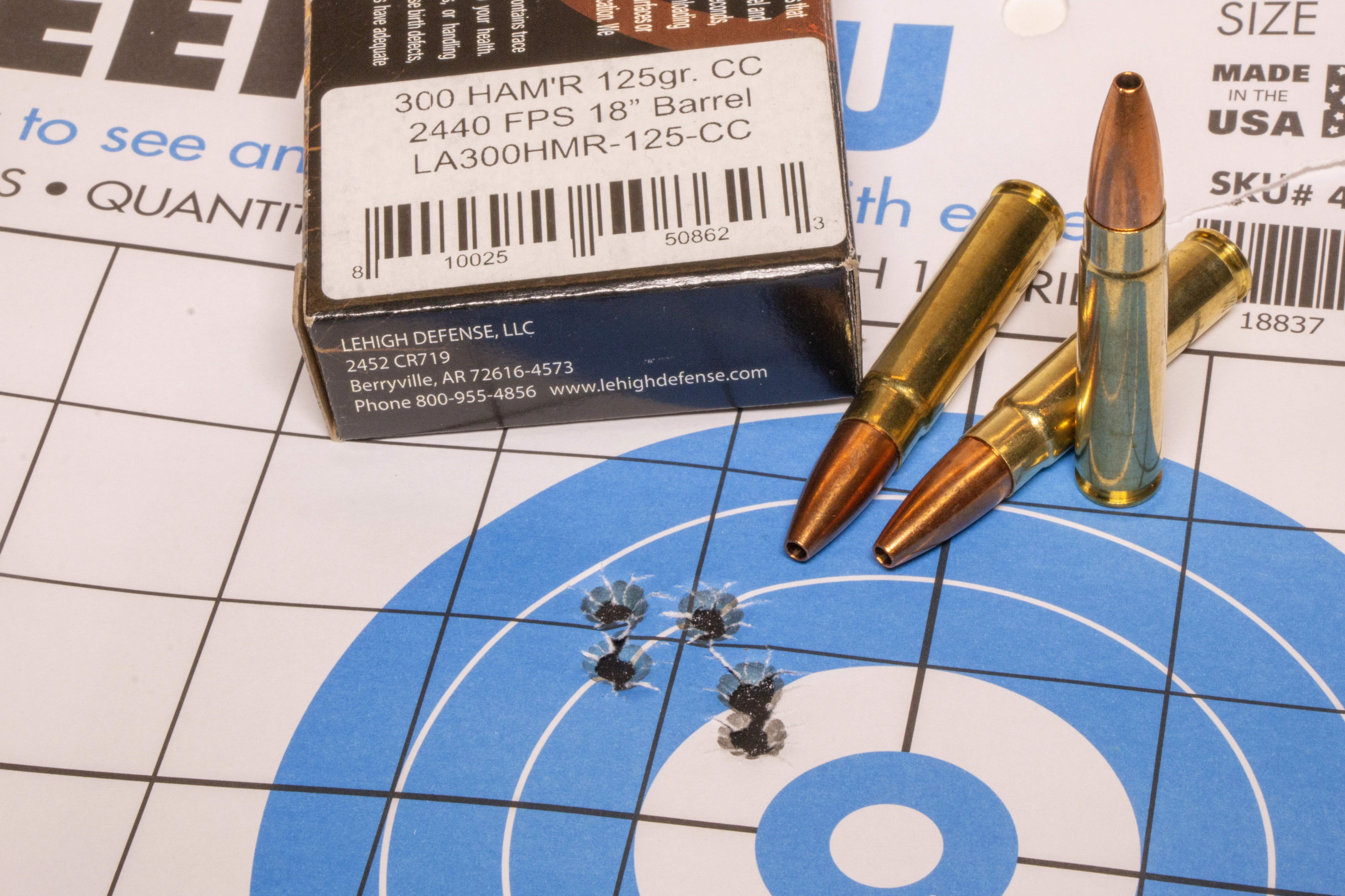 A rifle target showing five bullet holes in a group with a box of ammo and three loose cartridges.