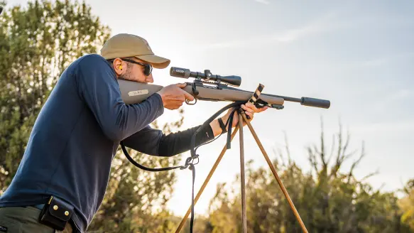 A shooting fires a bolt-action rifle resting on shooting sticks with trees and brush in background