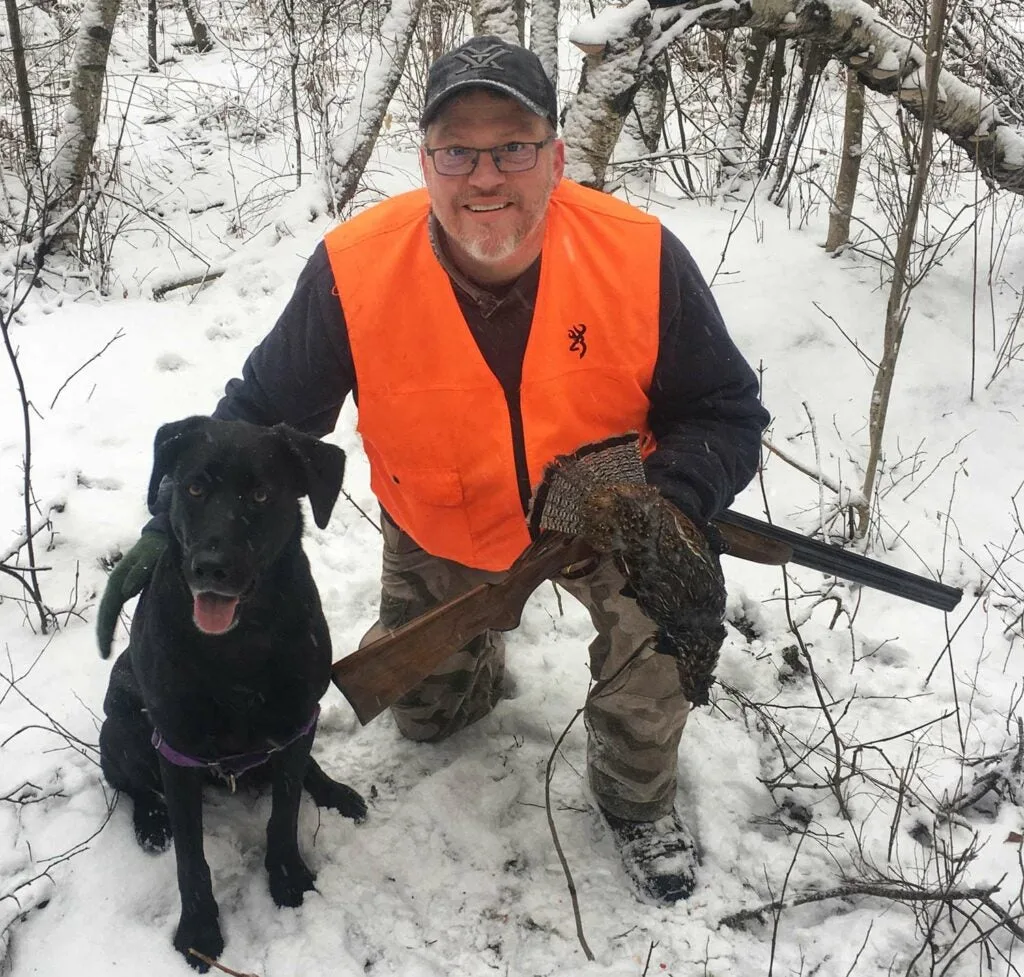 A hunter kneels in the snow and pets his hunting dog while holding a grouse.