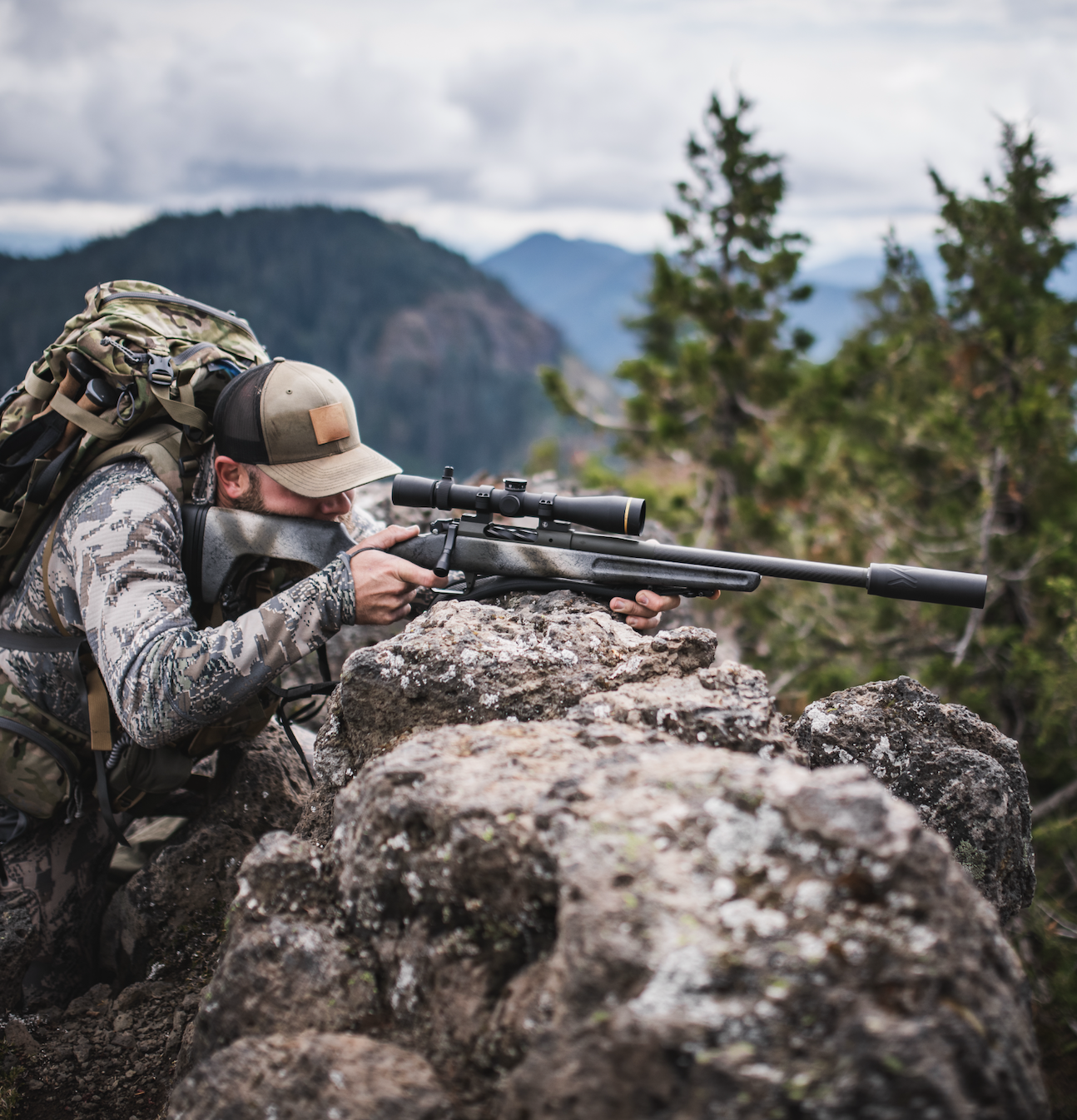A hunter shoots a rifle from a rock outcropping.