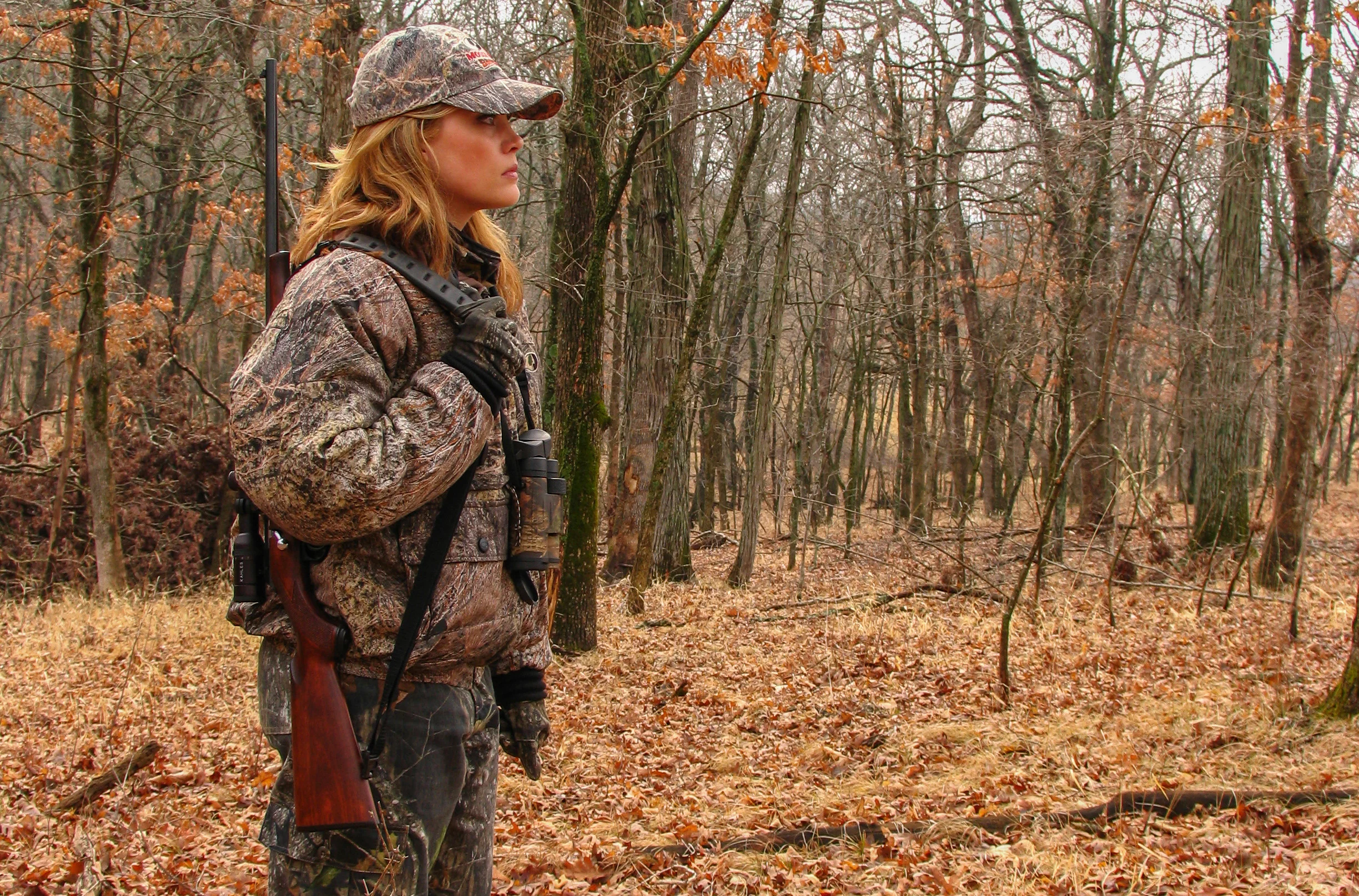 A female hunter walks through the woods with a rifle supported on her shoulder with a carry strap. 