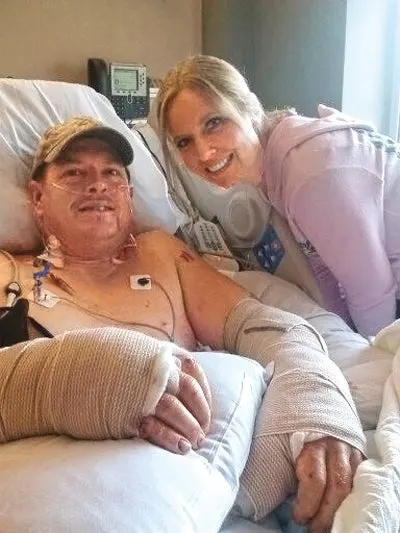 A man in a hat with bandaged arms rests in a hospital bed next to a woman.