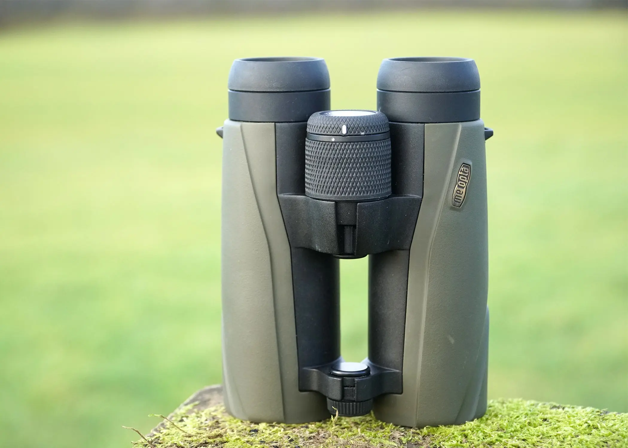 Meopta Meo Pro Air 10x42 binocular sitting on mossy wood with green field in background