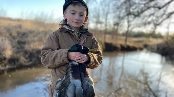 boy holds two ducks and stands in front of pond in lightly wooded area