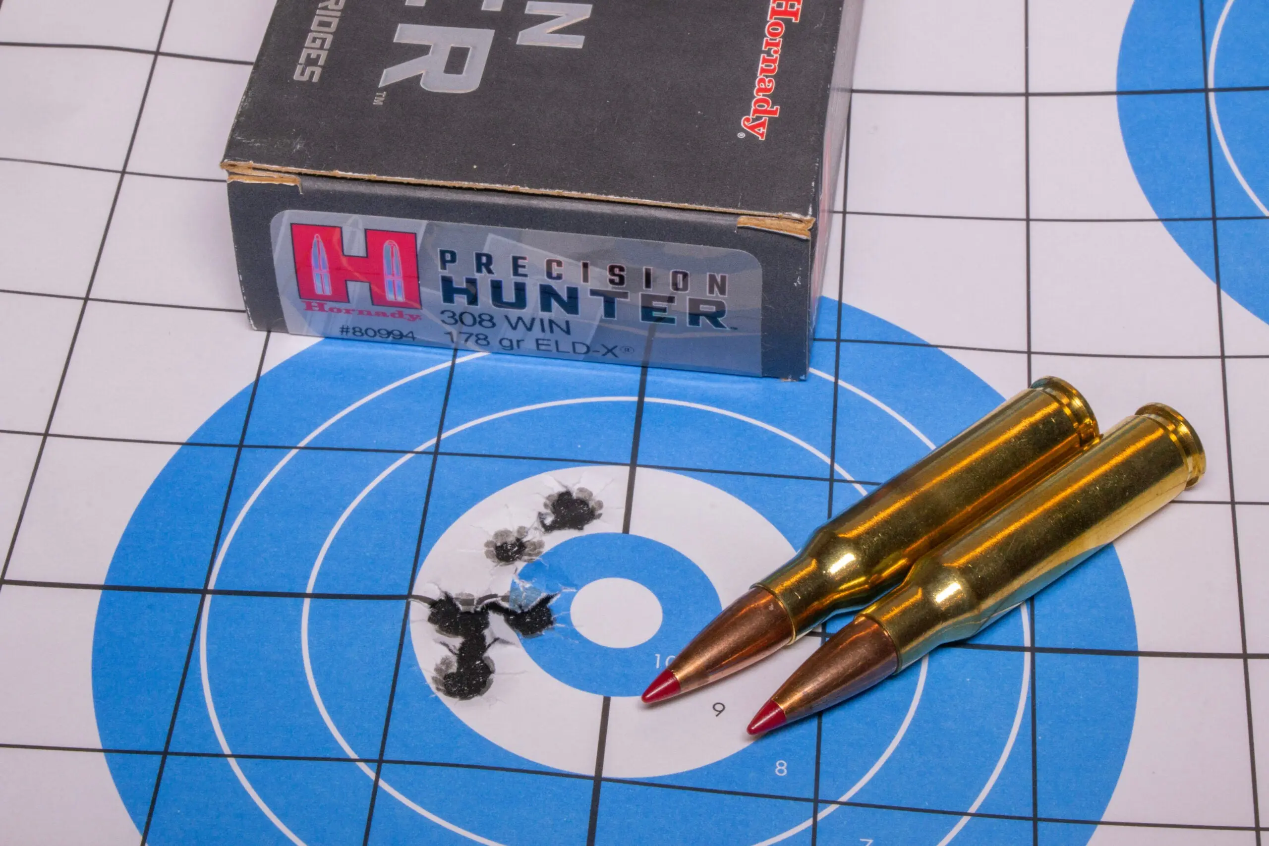 A rifle target showing five bullet holes in the center, with a box of ammo and two loose cartridges.