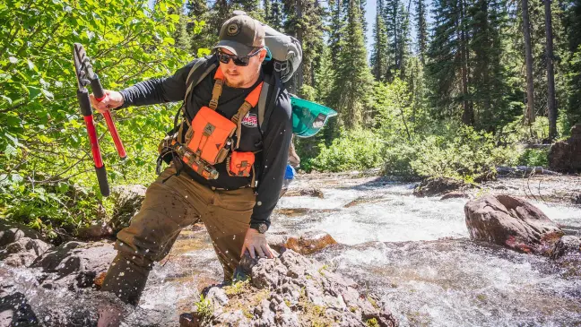 hiker-angler crosses stream holding brush clippers