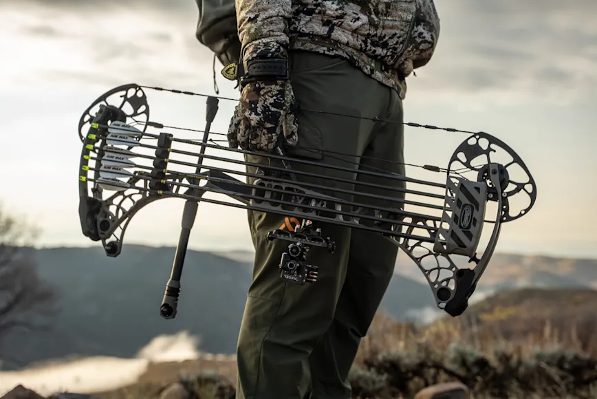 A bowhunter in a mountain landscape holds a compound bow by his side.