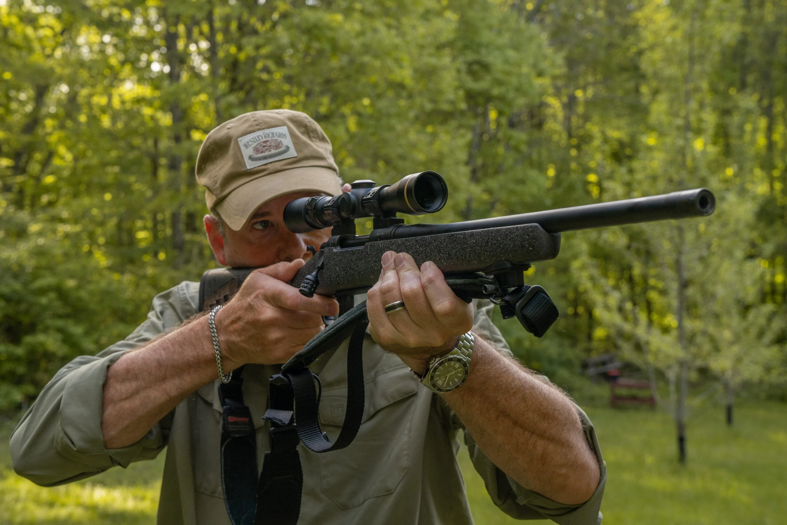 A shooter conducts dry-fire practice with a bolt-action rifle with green woods in background