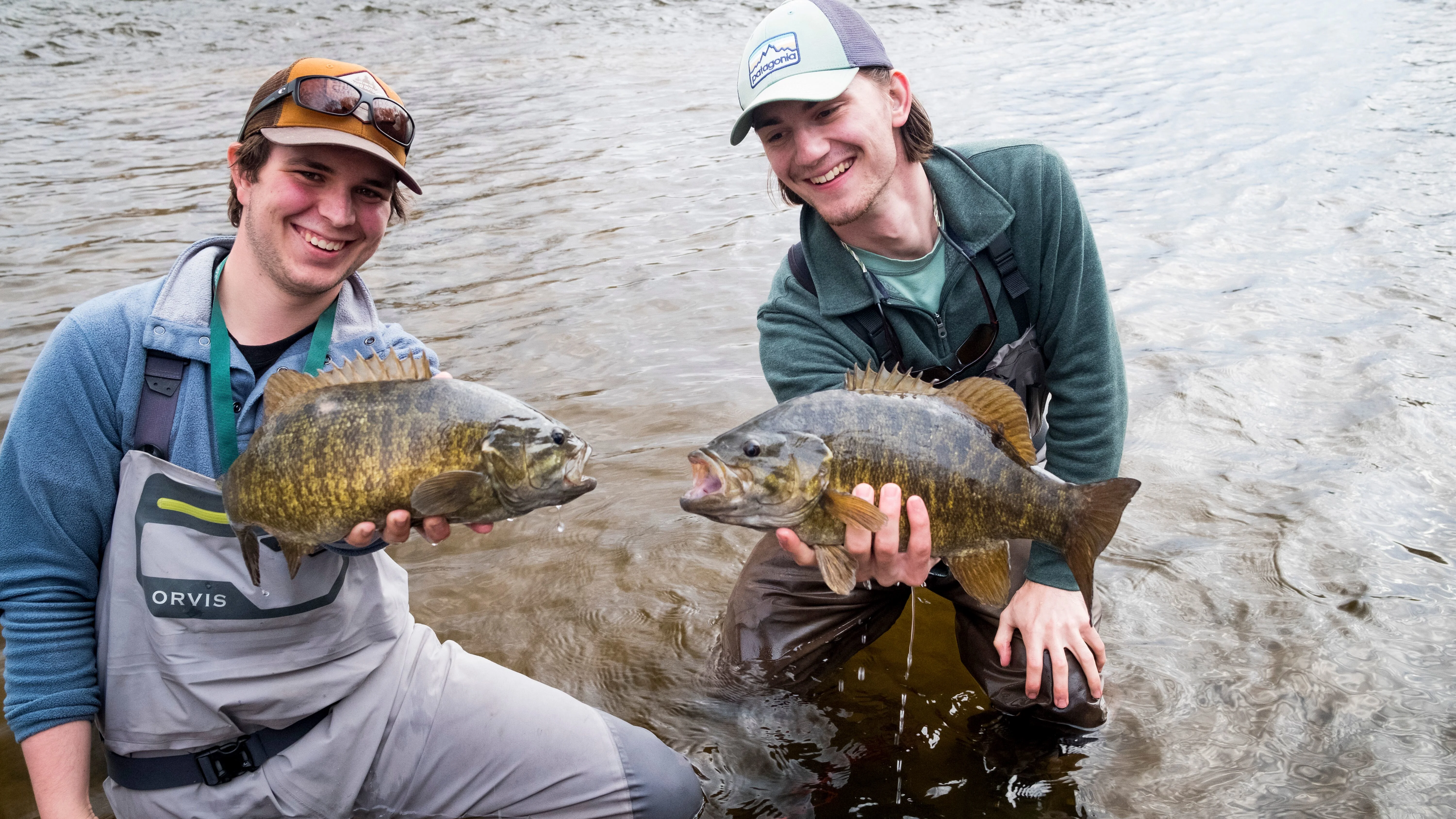 Two fishermen hold up smallmouth bass while kneeling in a river