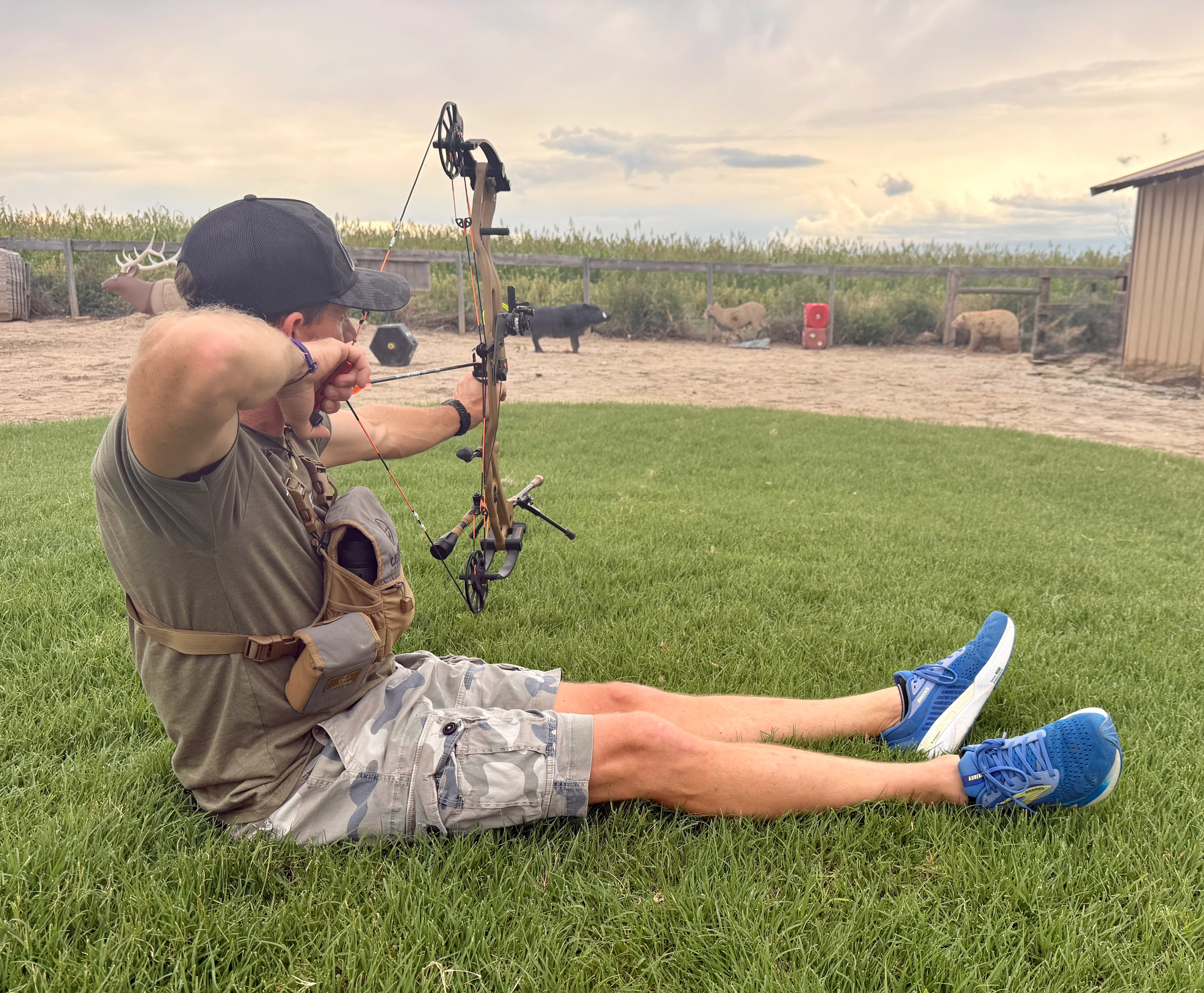 An archer draws his bow while sitting on the ground on an archery range.
