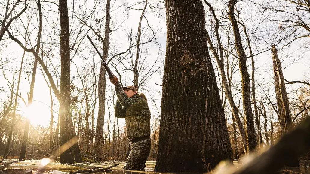 duck hunter standing in flooded timber with his gun pointed toward the sky