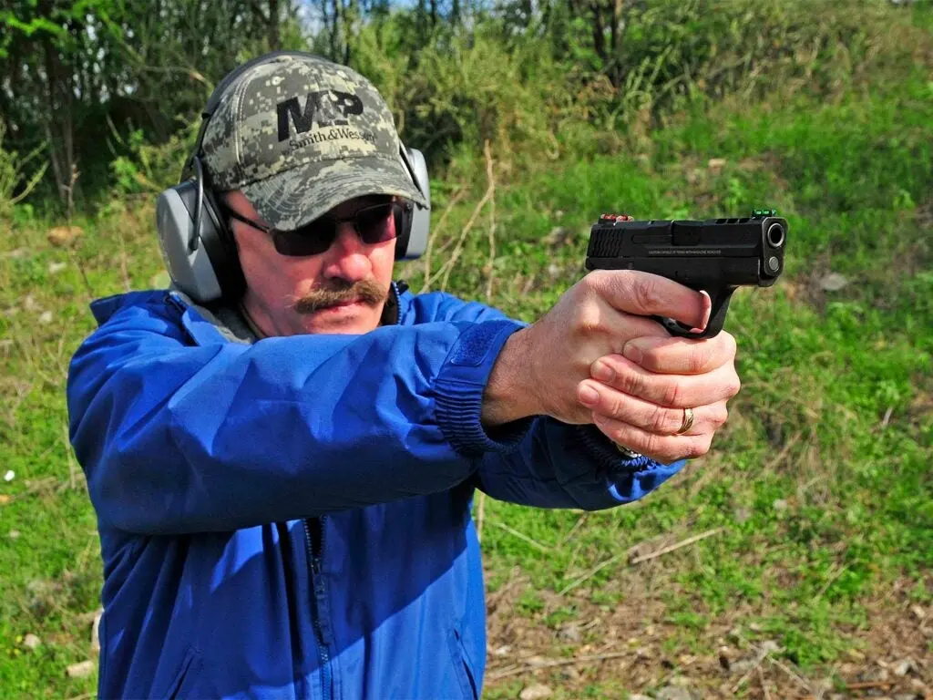 A man in a blue jacket aims a handgun in the woods.