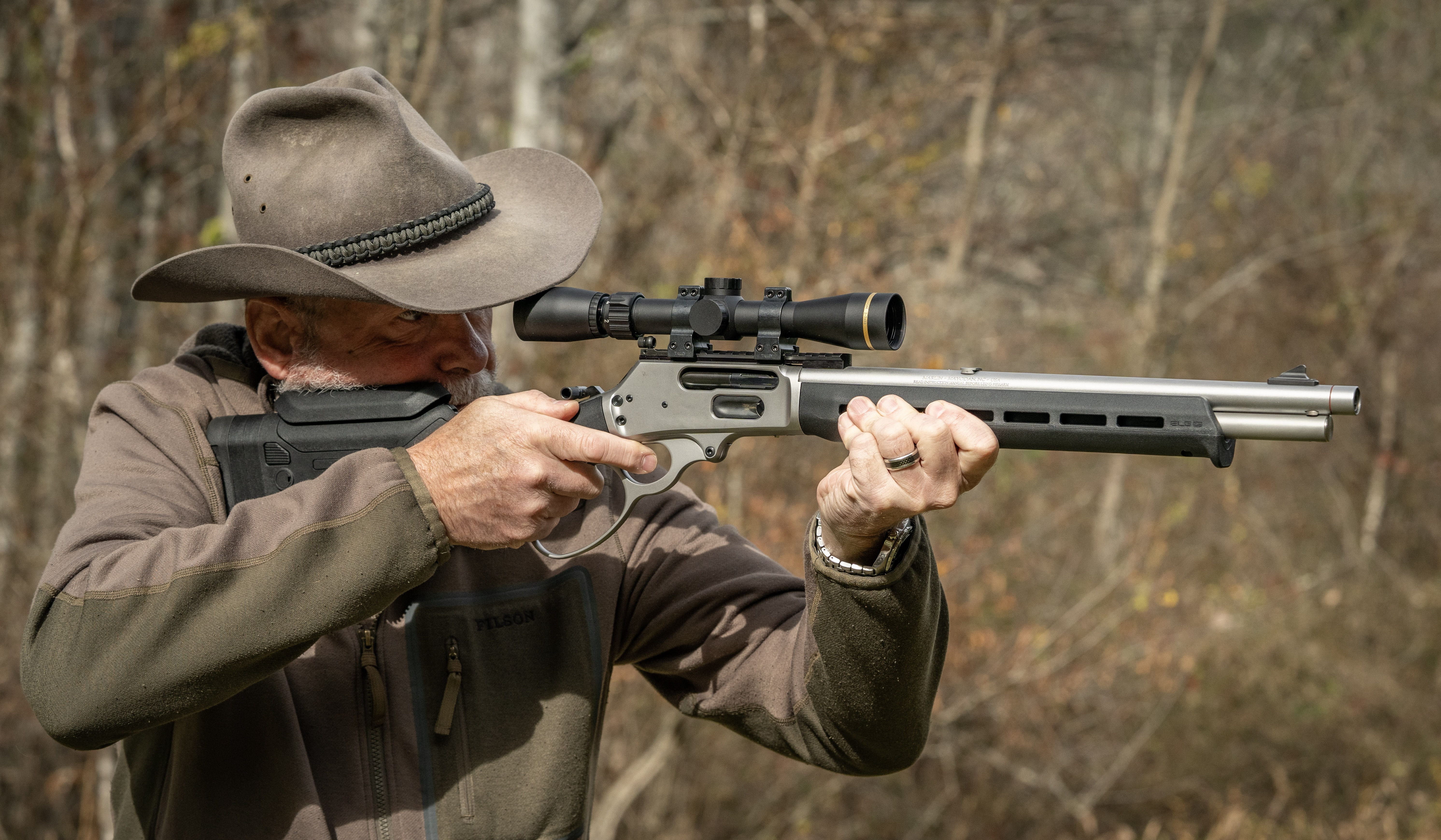 A shooter fires a lever action rifle with woods in the background. 