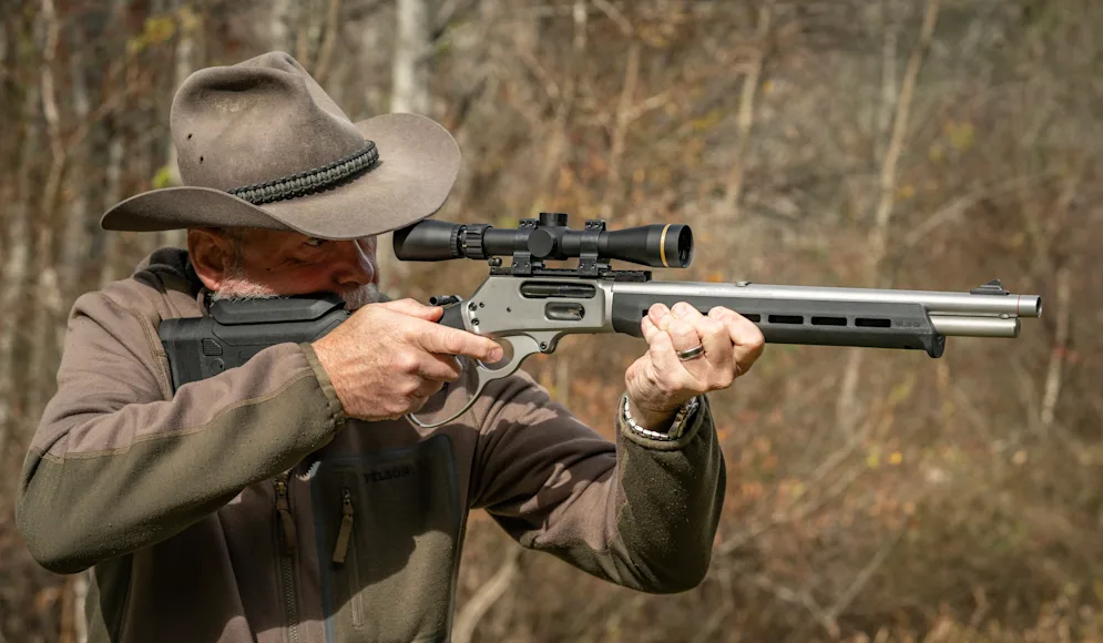 A shooter fires a lever action rifle with woods in the background.