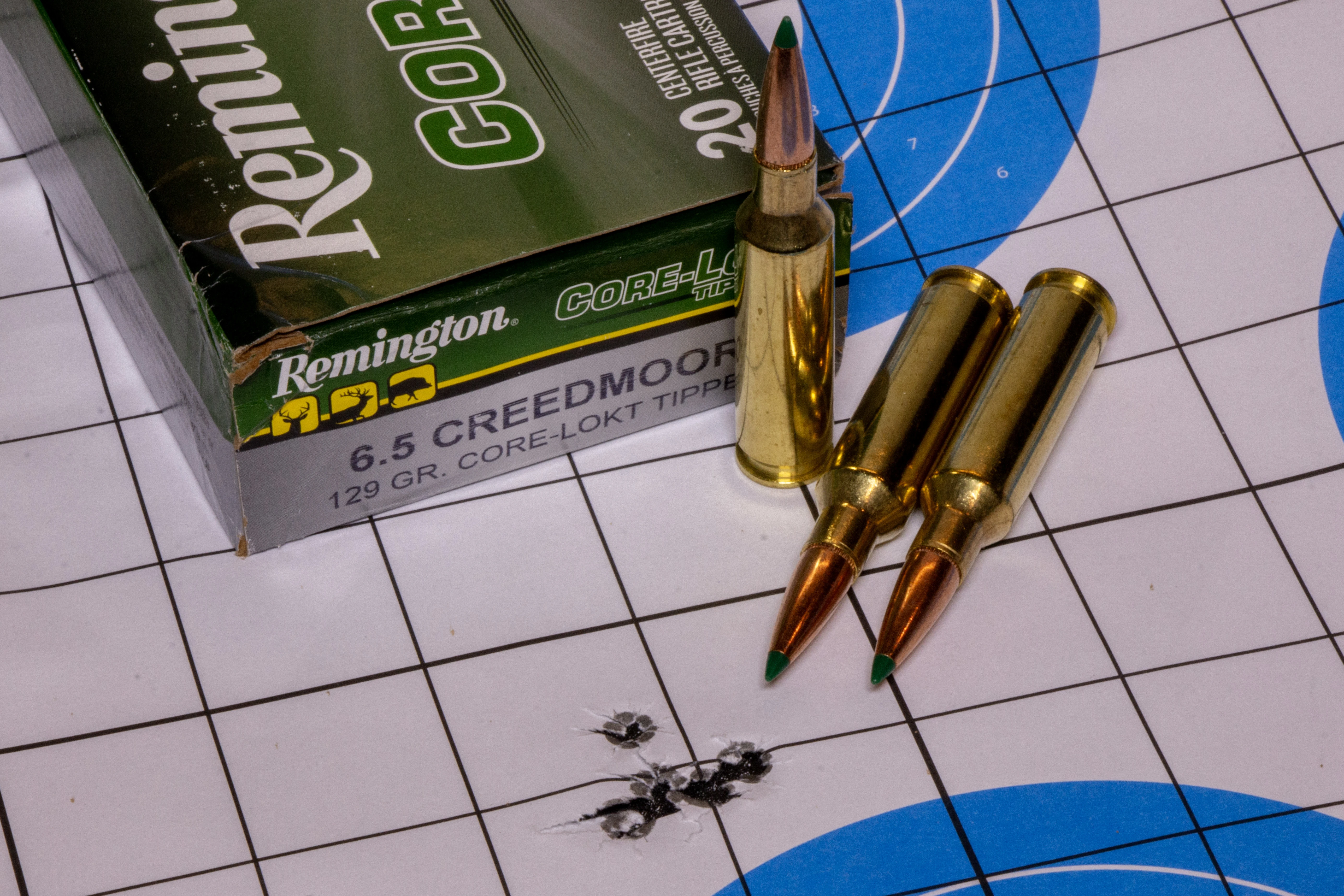 A rifle target with five bullet holes in it, with a box of ammo and three loose cartridges nearby.