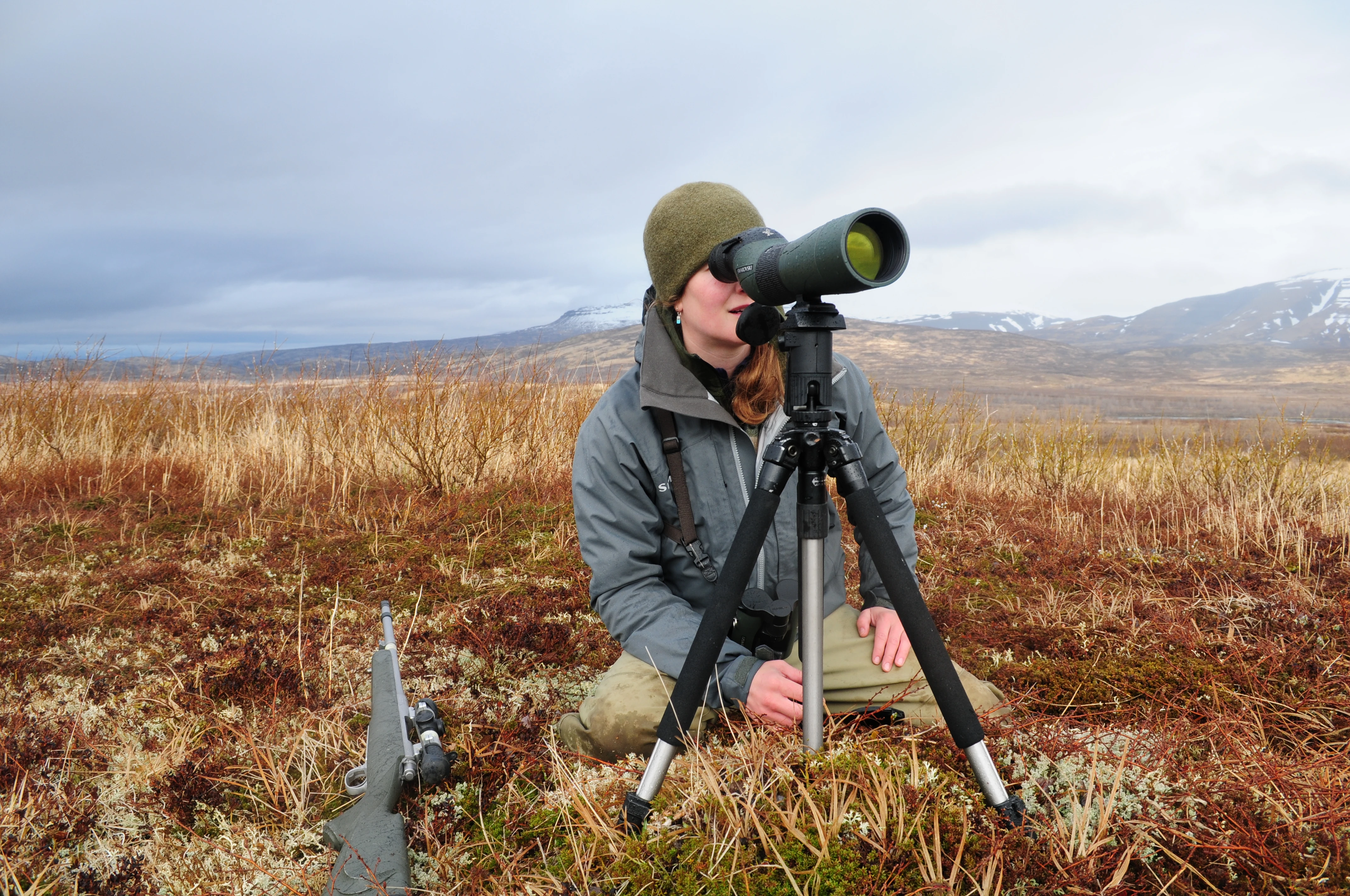 Alaska bear guide Tia Shoemaker sits on the ground with rifle handy and looks for game through a spotting scope.