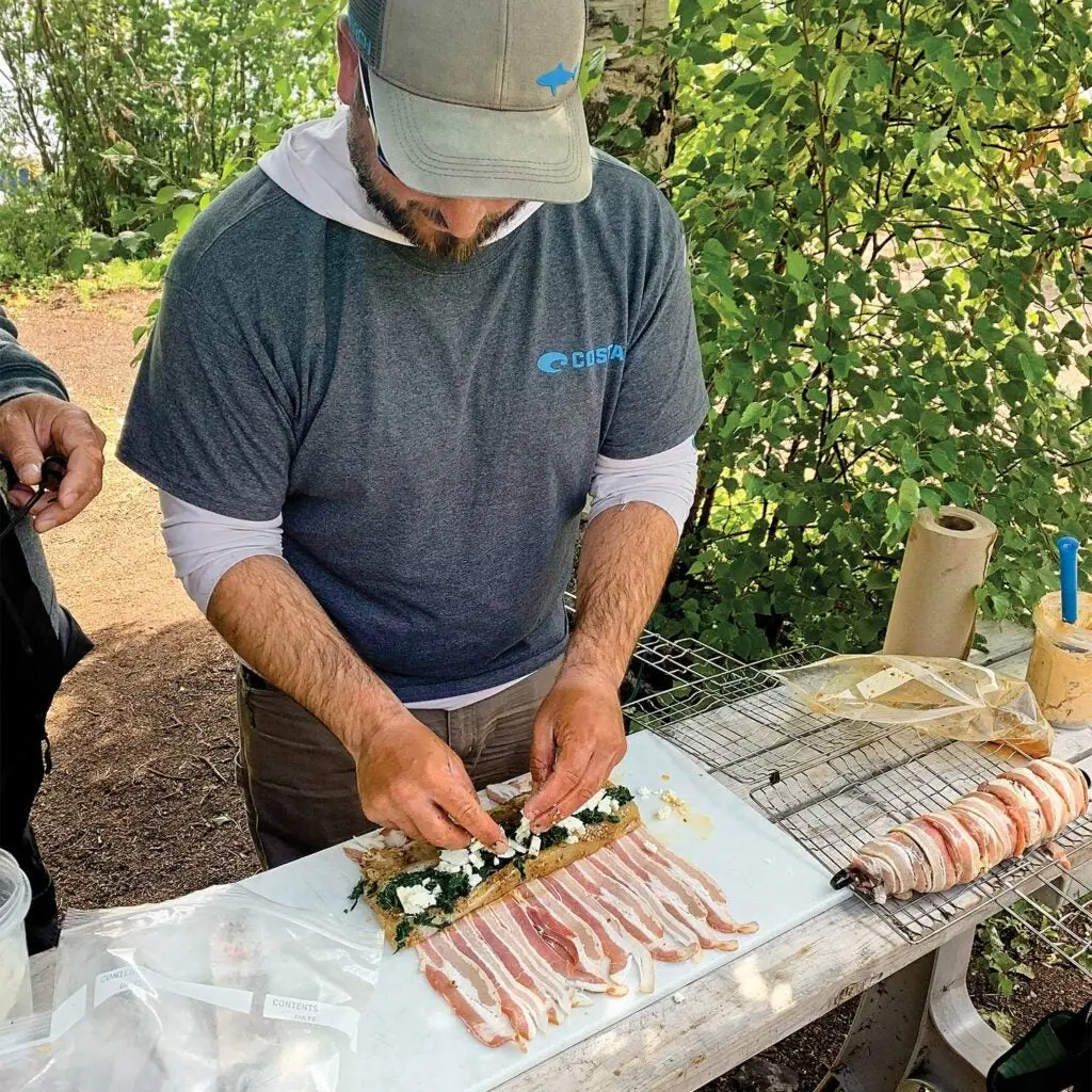 A bearded man wearing a hat prepares northern pike meat with toppings before wrapping the fillings in strips of bacon.