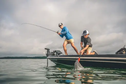 A fisherman fights a bass while a another boat passenger weilds the net. 