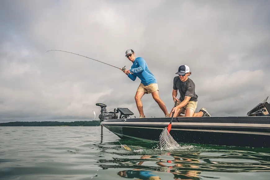 A fisherman fights a bass while a another boat passenger weilds the net.