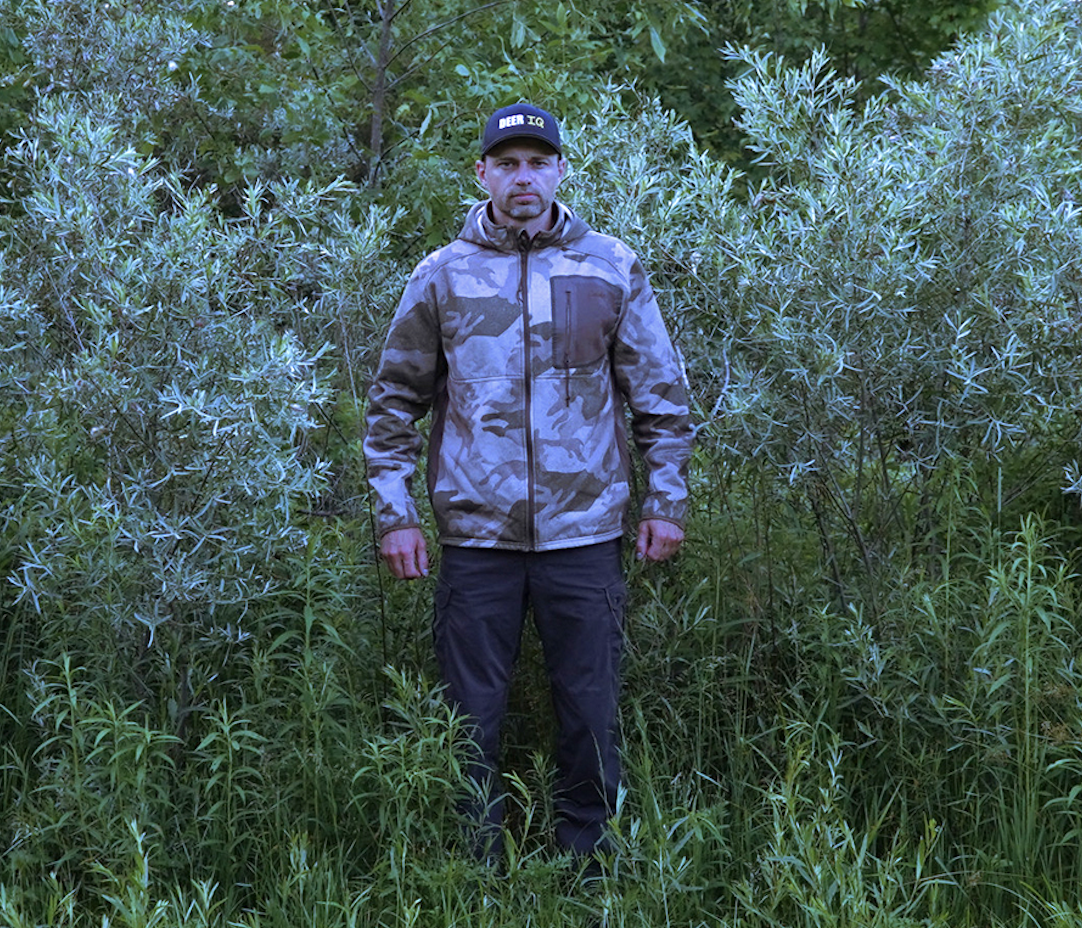 A hunter wearing a camo jacket stands in green, leafy bushes.