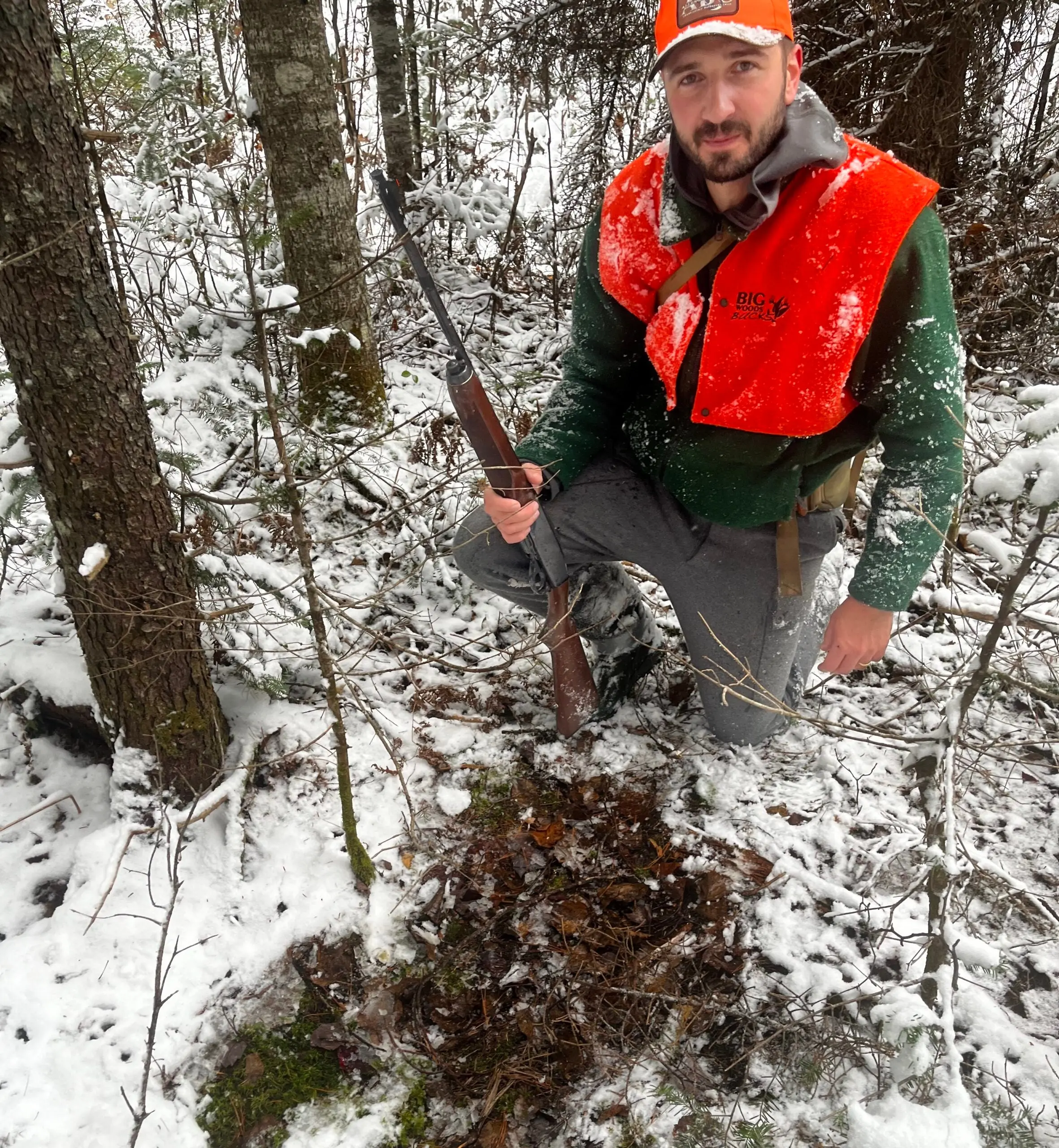 A hunter with a rifle kneels next to a deer bed in the snow