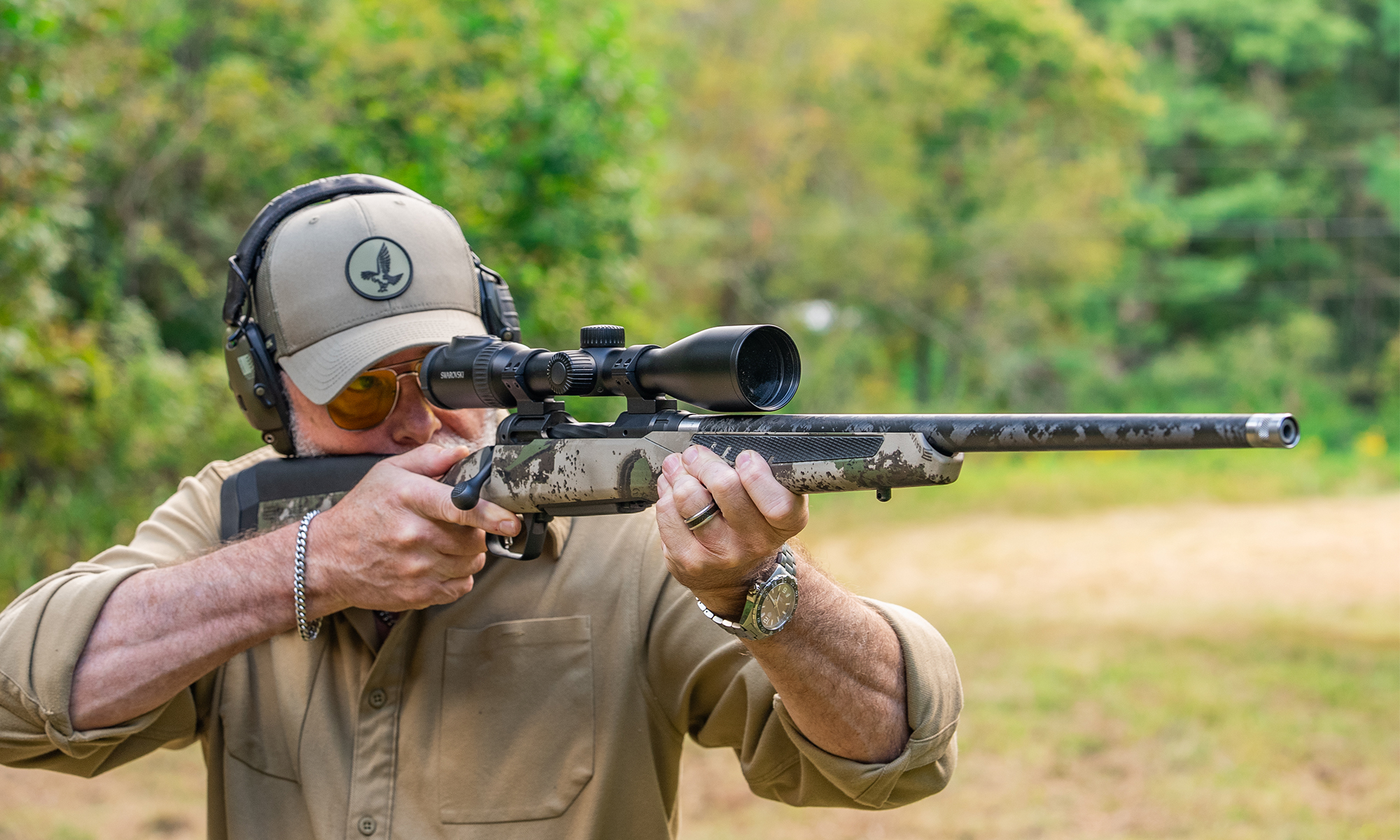 A shooter fires a bolt-action rifles in a field with trees in background. 