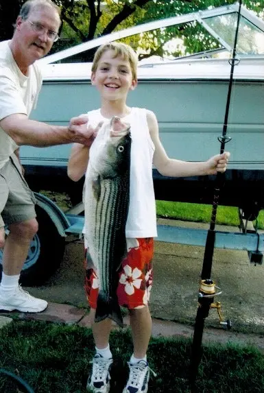 A boy in a white shirt and red shorts holds a striped bass in front of a boat