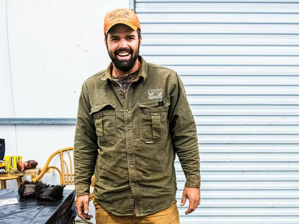 A bearded man in a cap smiles at the camera while standing next to a wooden table.