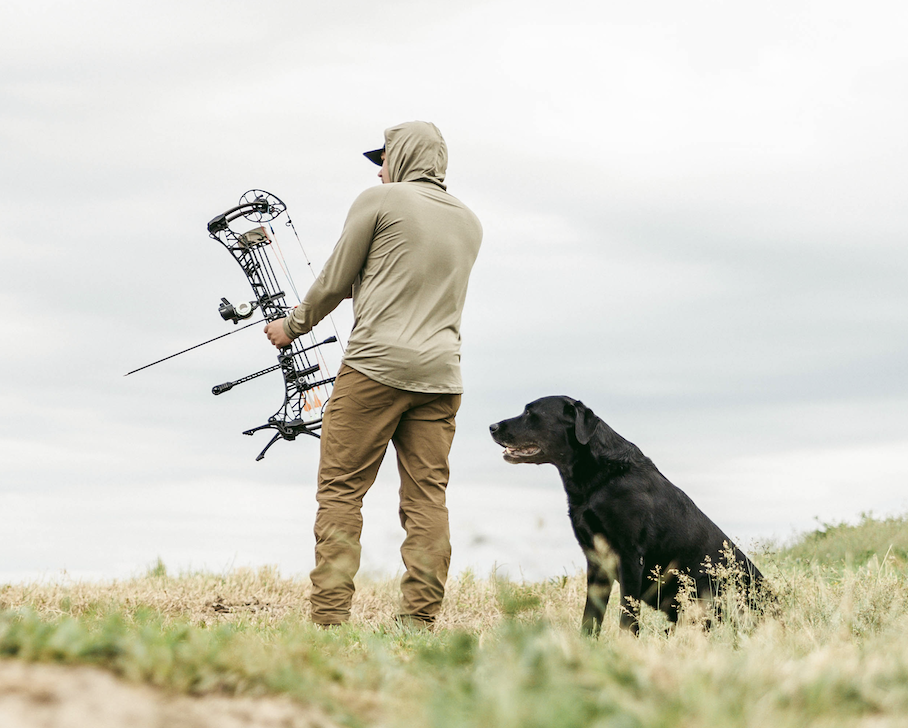A bowhunter shoots at a target in a field with his dog standing behind him. 