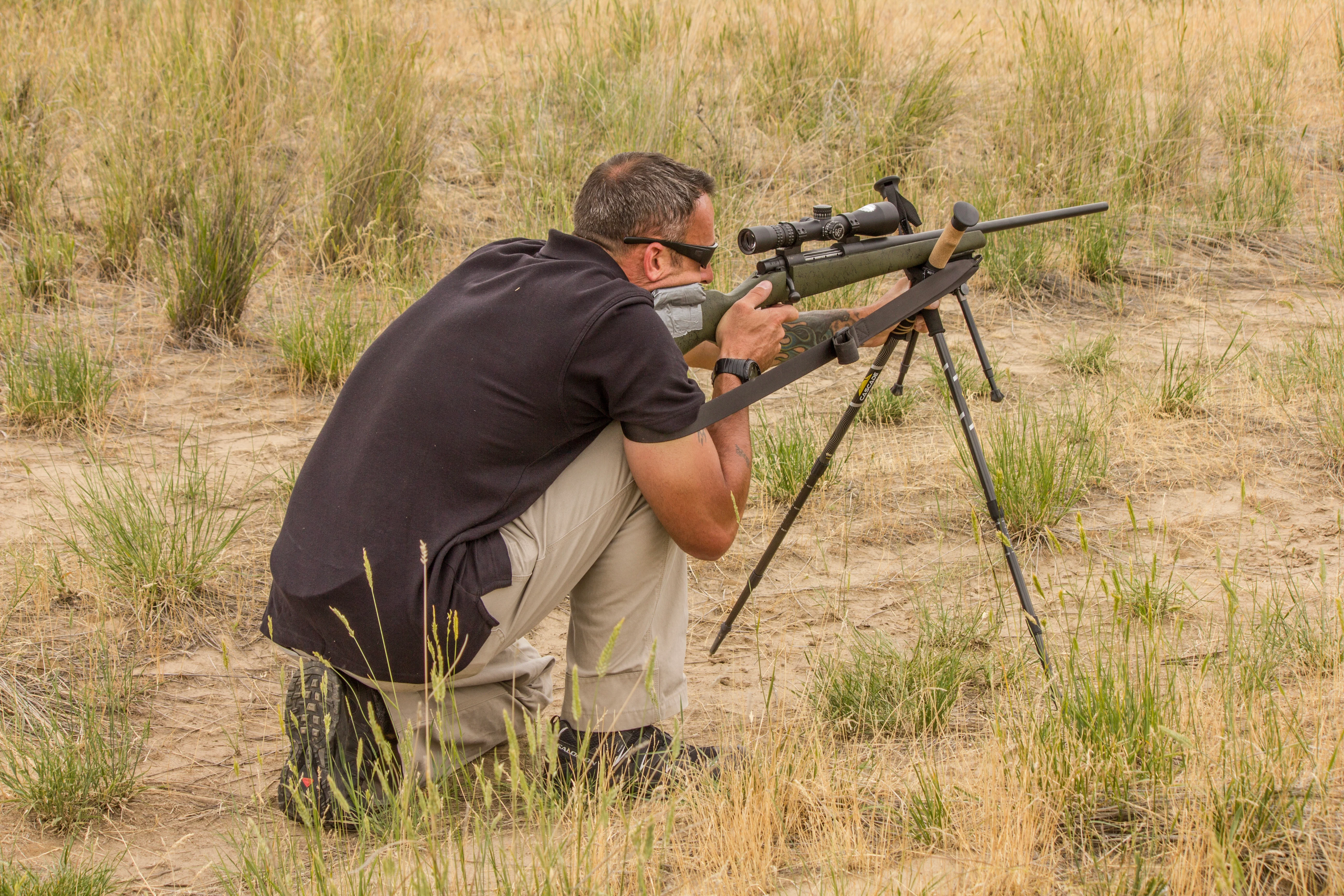 A shooter fires a rifle from a kneeling position with shooting sticks and sling for added support. 