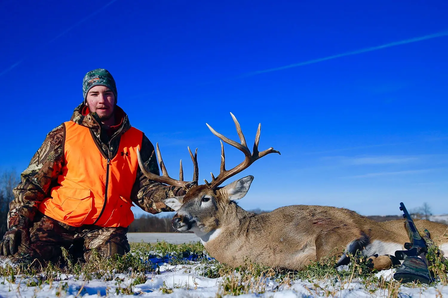 hunter in orange vest and camo kneels next to large whitetail buck, with some snow on the ground