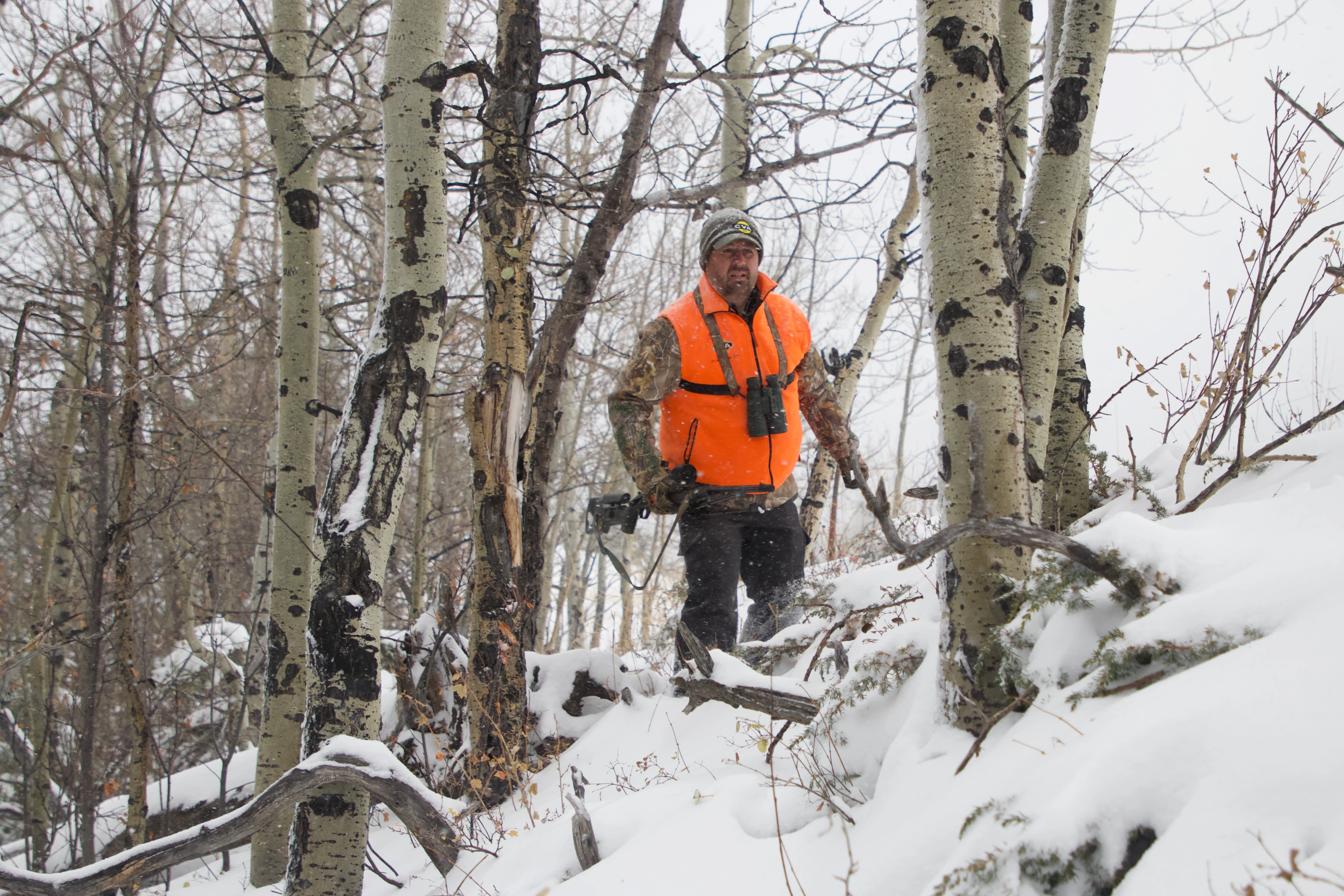 A hunter weather blaze orange still-hunts a wooded slope with a rifle. 