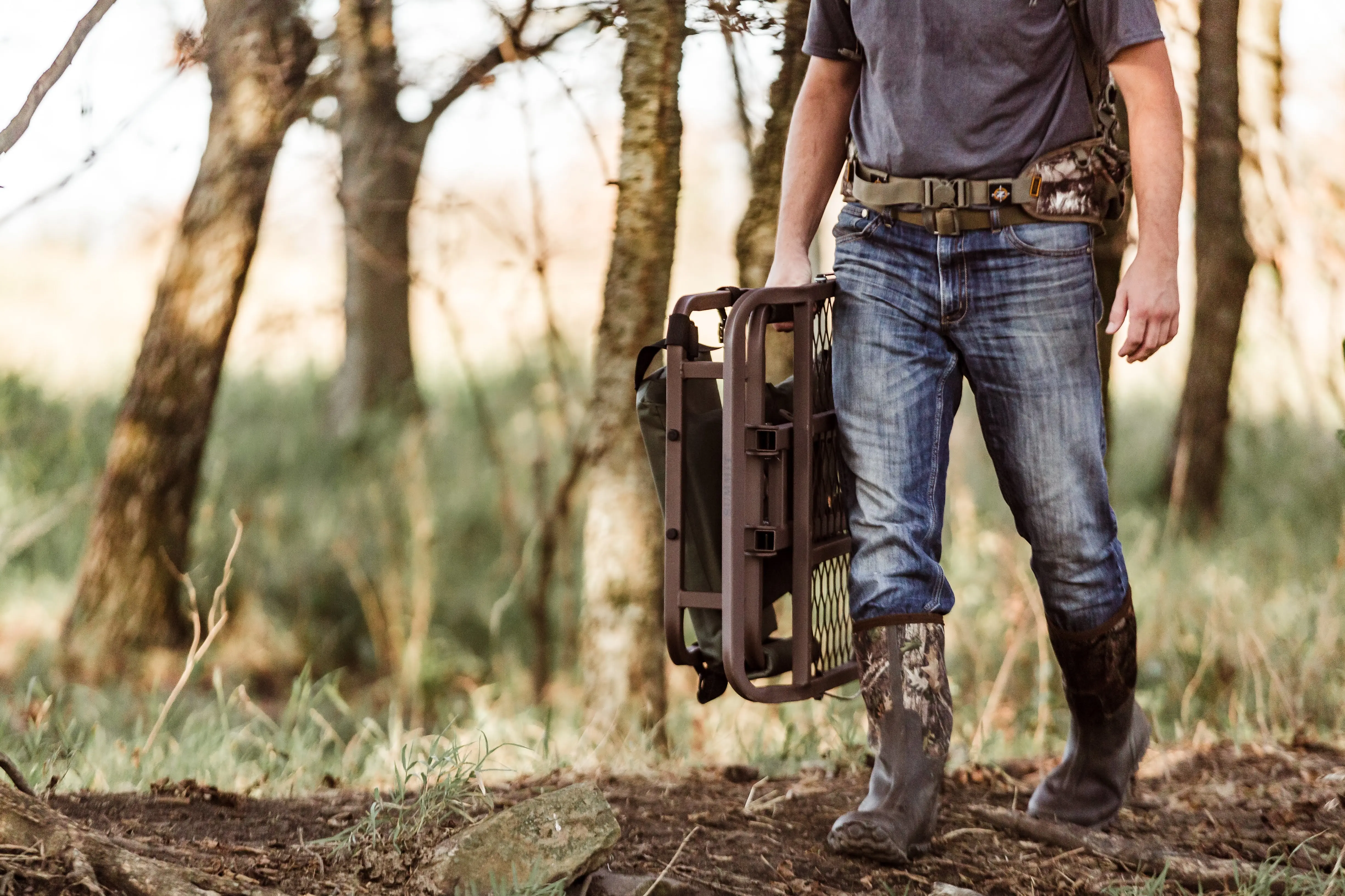 A hunter in jeans and a tee shirt carries a hang-on tree stand into the woods. 