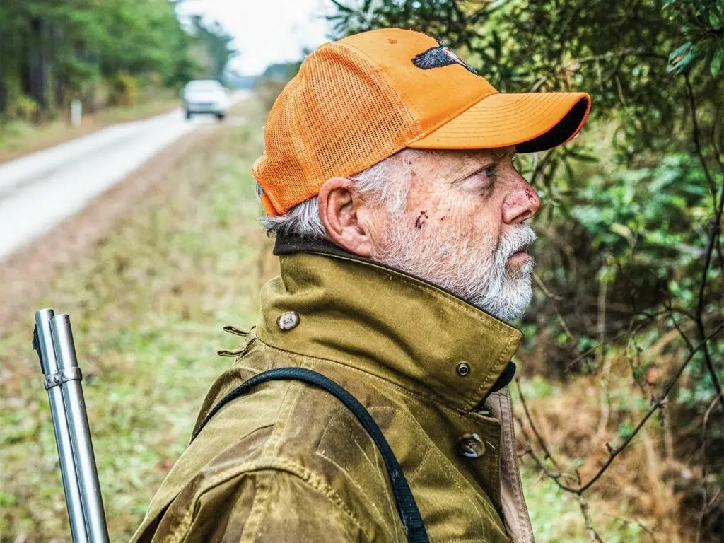 A man with a rifle slung over his shoulder and wearing an orange hunting cap looks into the treeline next to a road.