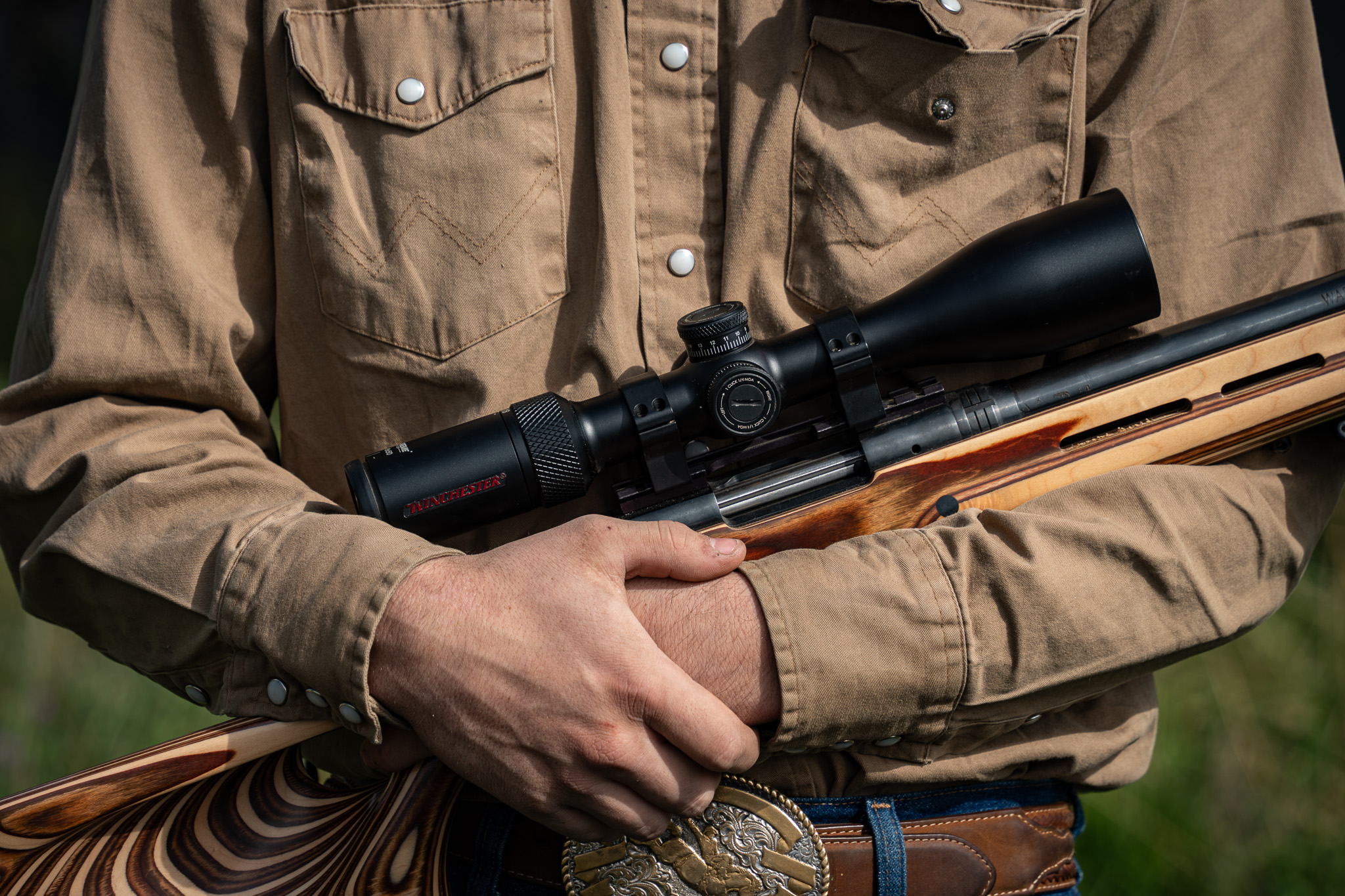 A man in a tan shirt holds a scoped rifle in his arms.