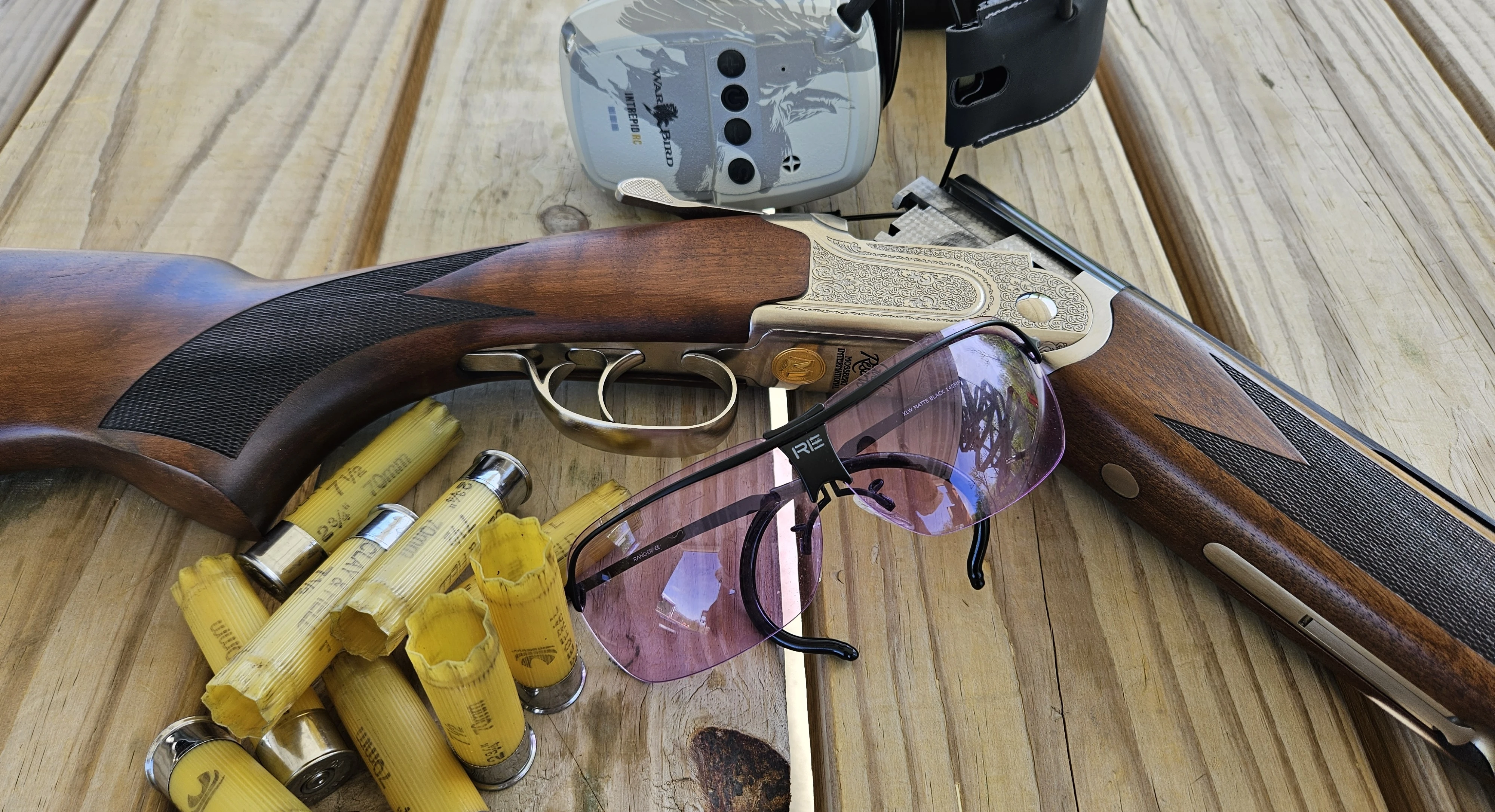 The new Mossberg Gold Reserve shotgun on a wood surface with ear muffs, shooting glasses, and empty shells. 