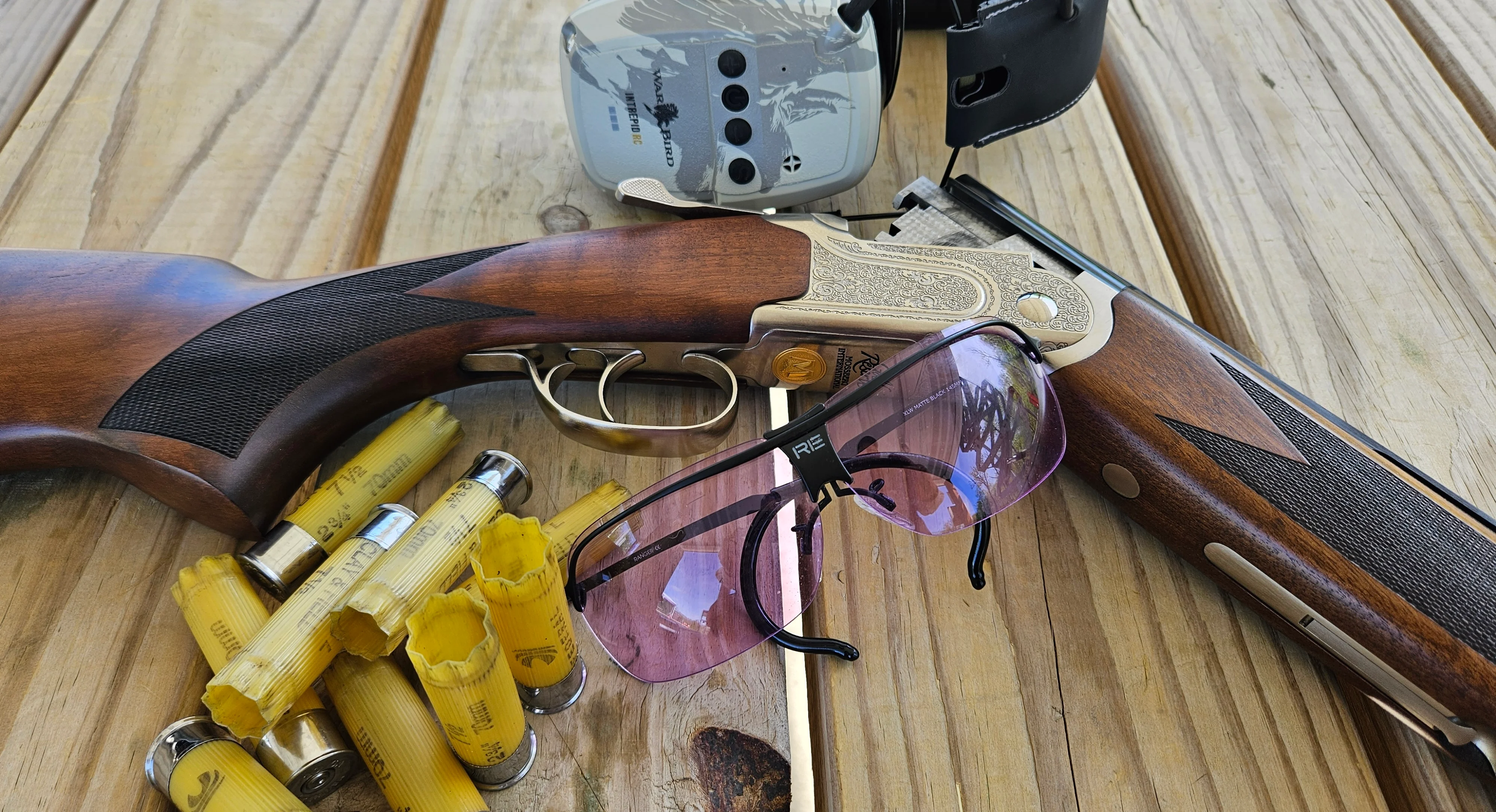 The new Mossberg Gold Reserve shotgun on a wood surface with ear muffs, shooting glasses, and empty shells. 