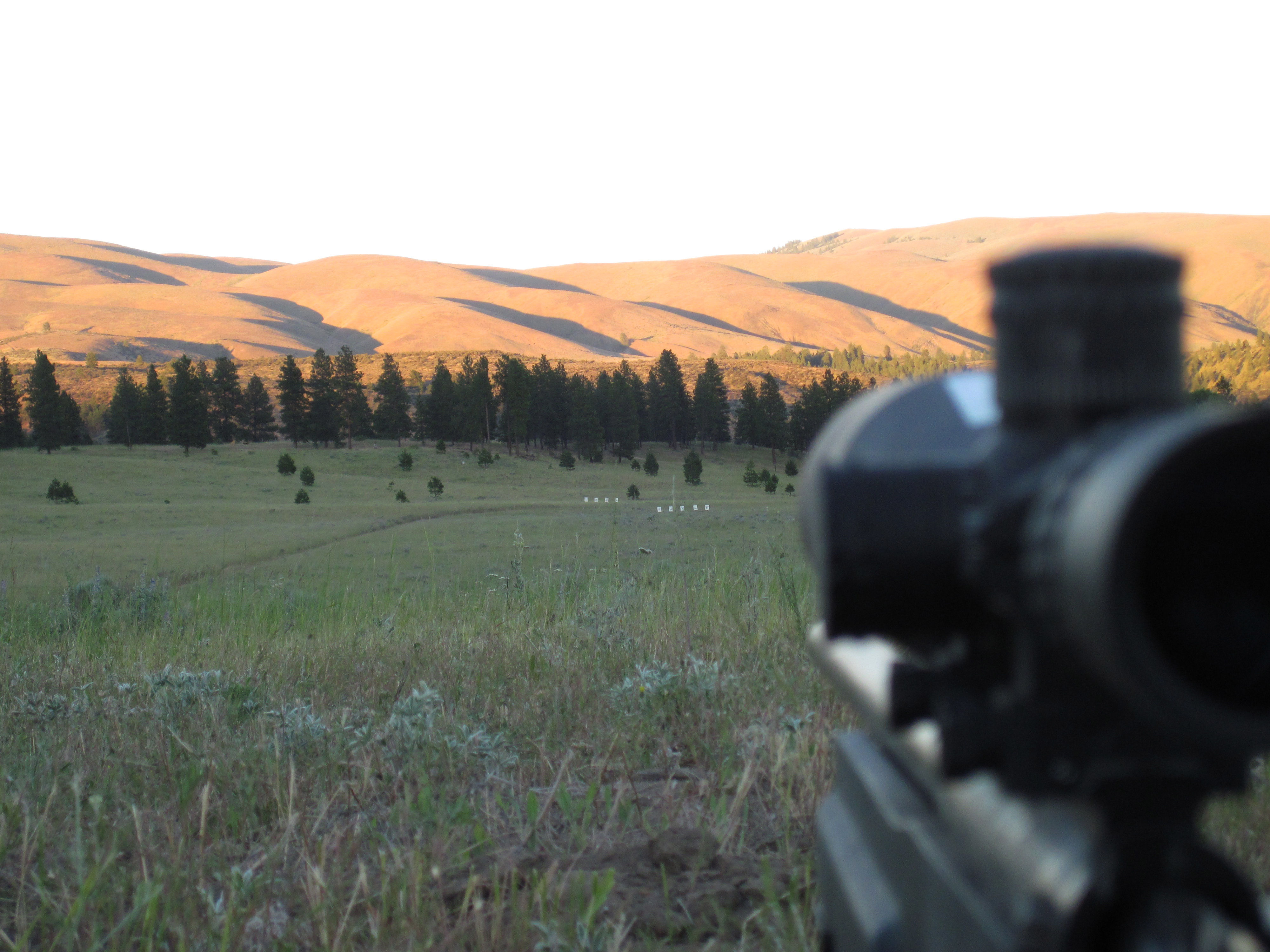 A riflescope in the foreground looks toward a row of steel targets off in the distance with mountains in background. 