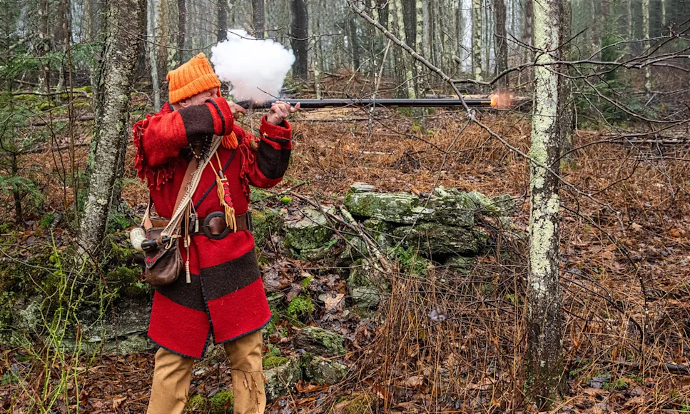 Man in traditional red jacket fires a flintlock rifle, with smoke billowing from the lock.