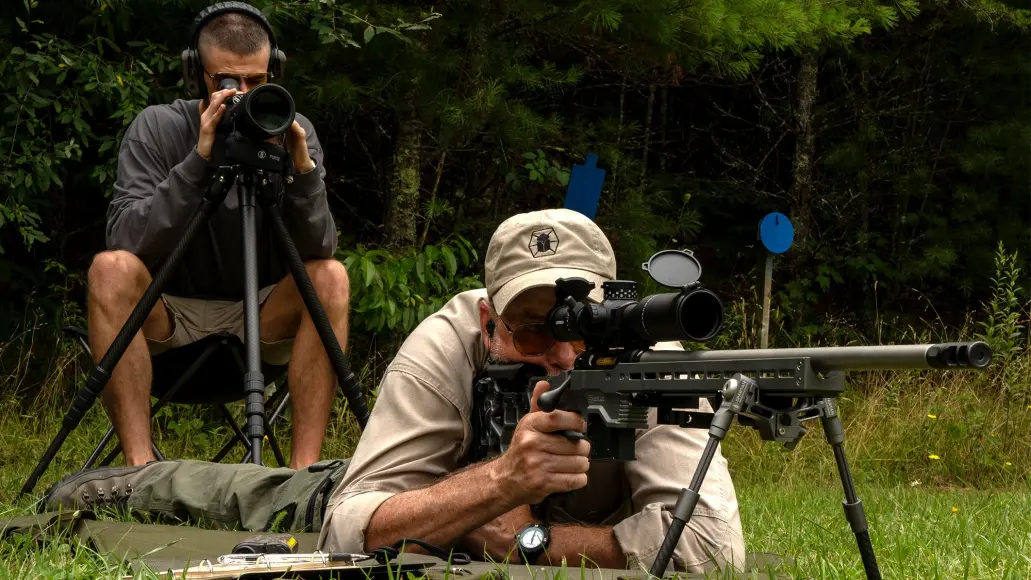 A man laying on the ground shooting a rifle while another looks through a spotting scope