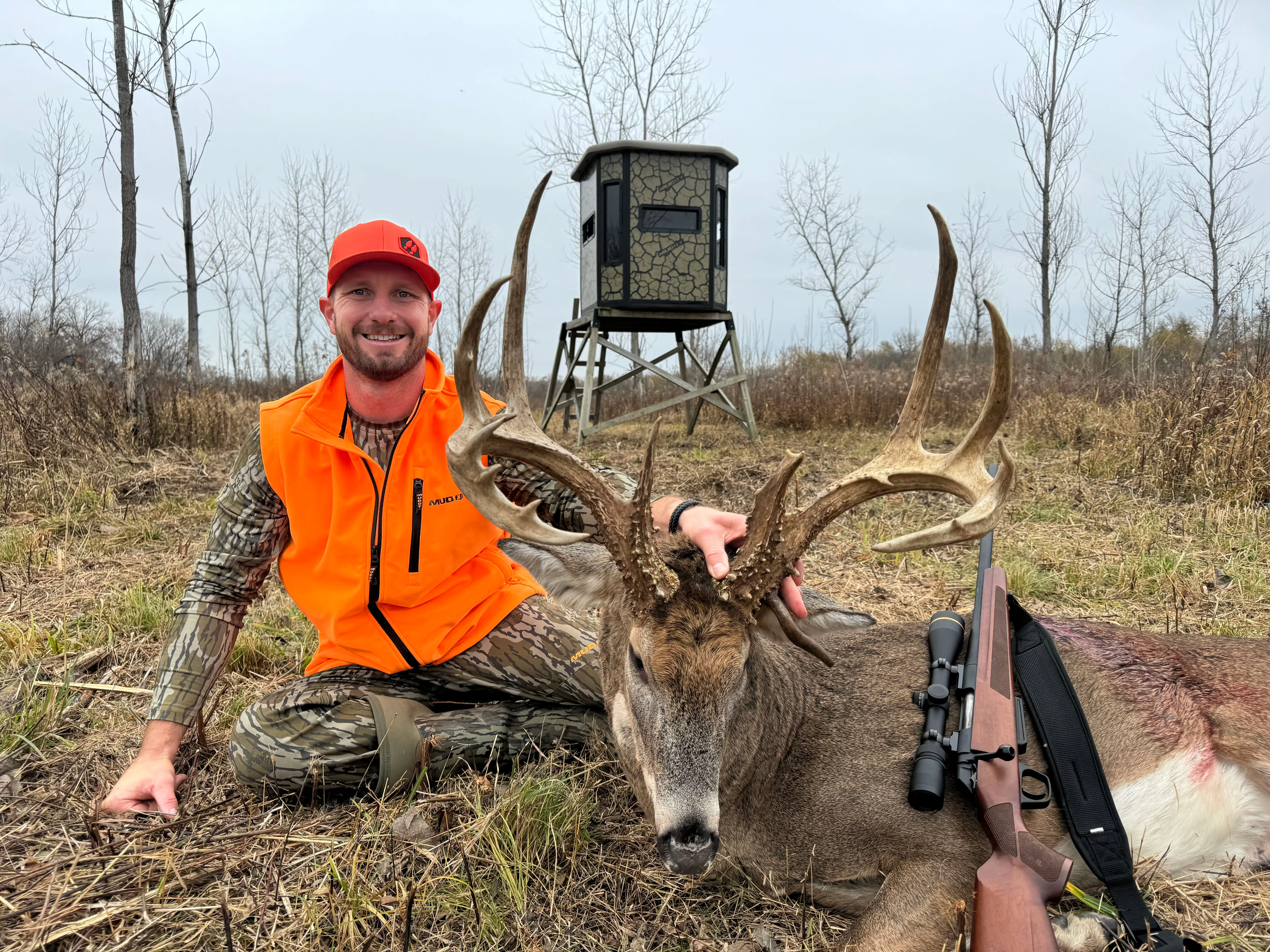 A hunter in orange poses in a field with a trophy whitetial buck he took with a rifle.