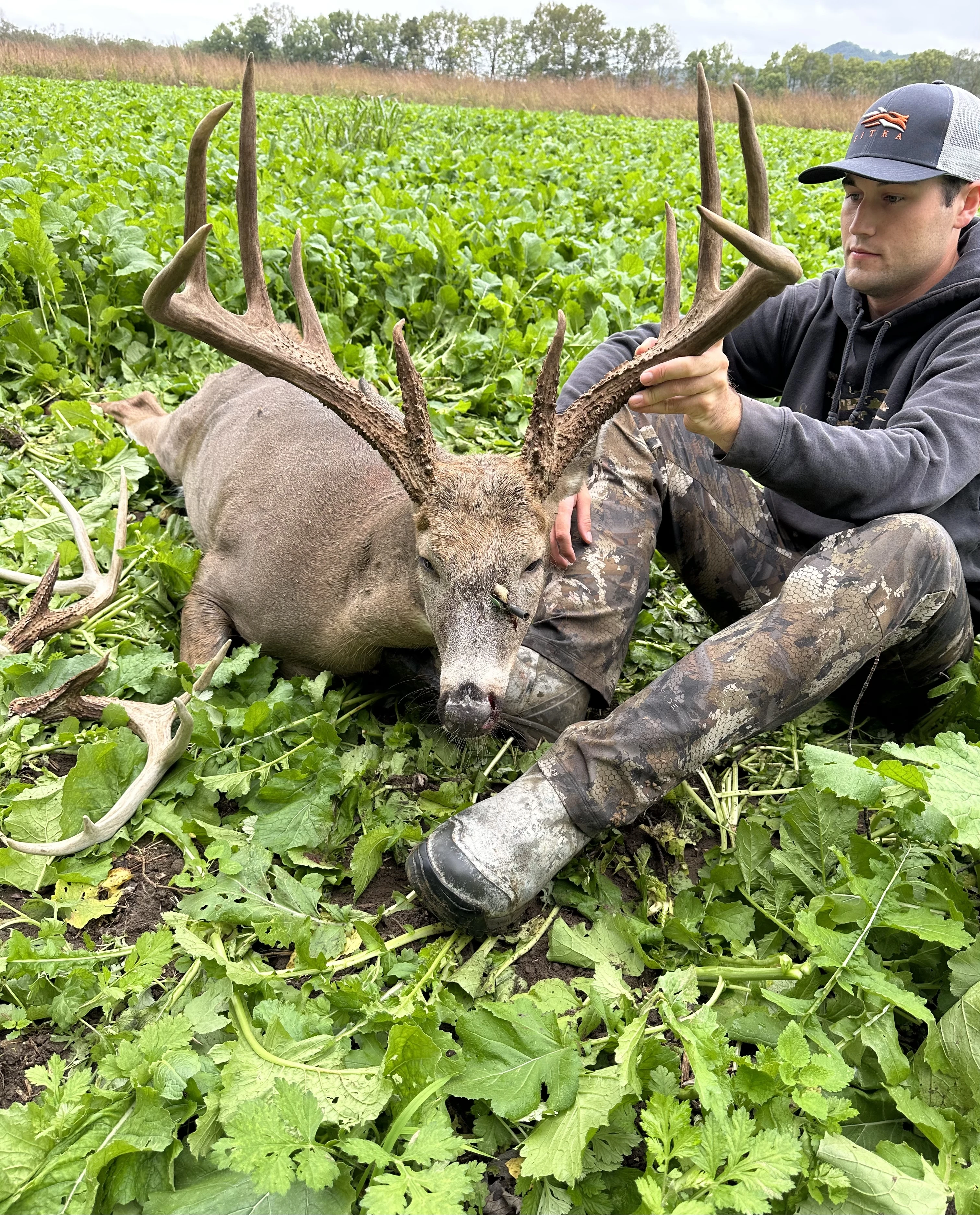 Ohio hunter Noah Dresbach sits in a green field and shows off a big whitetail buck he took with a bowl. 