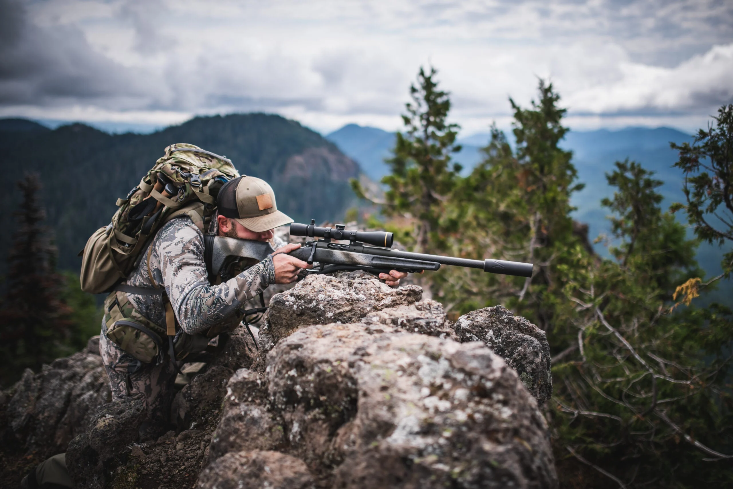 A hunter shoots a rifle from a rock outcropping on a mountain