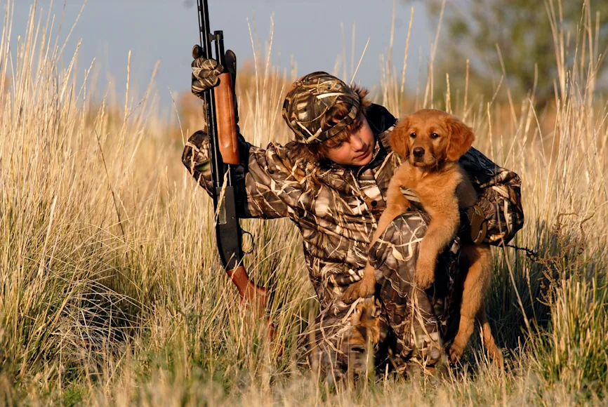 A hunter and a hunting dog duck hunting by a pond.